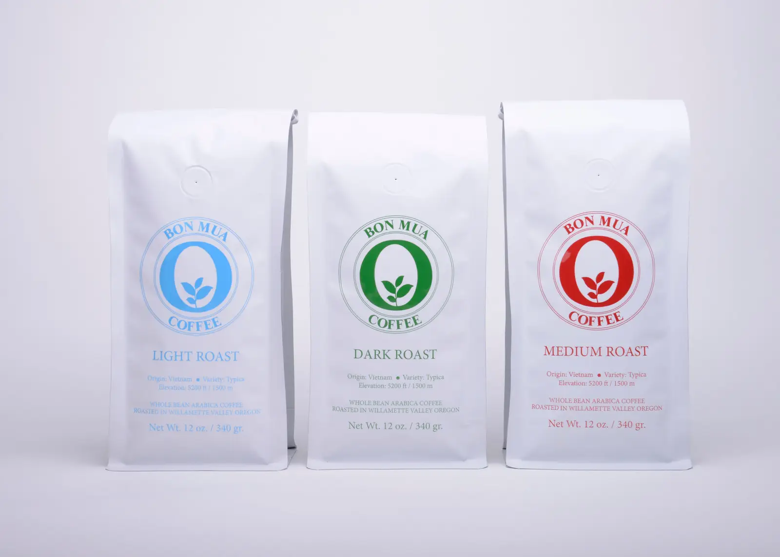 Three white Bon Mua coffee bags standing together — a blue-logo Light Roast, a green-logo Dark Roast, and a red-logo Medium Roast — on a pale grey background.