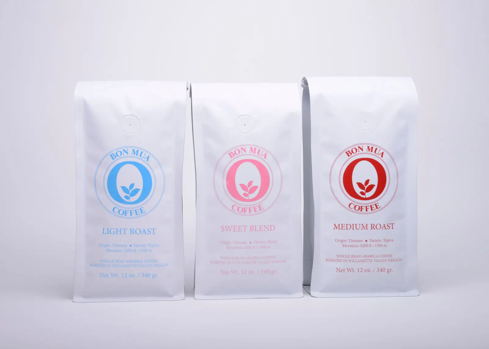Three white Bon Mua coffee bags standing together — a blue-logo Light Roast, a pink-logo Sweet Blend, and a red-logo Medium Roast — on a pale grey background.