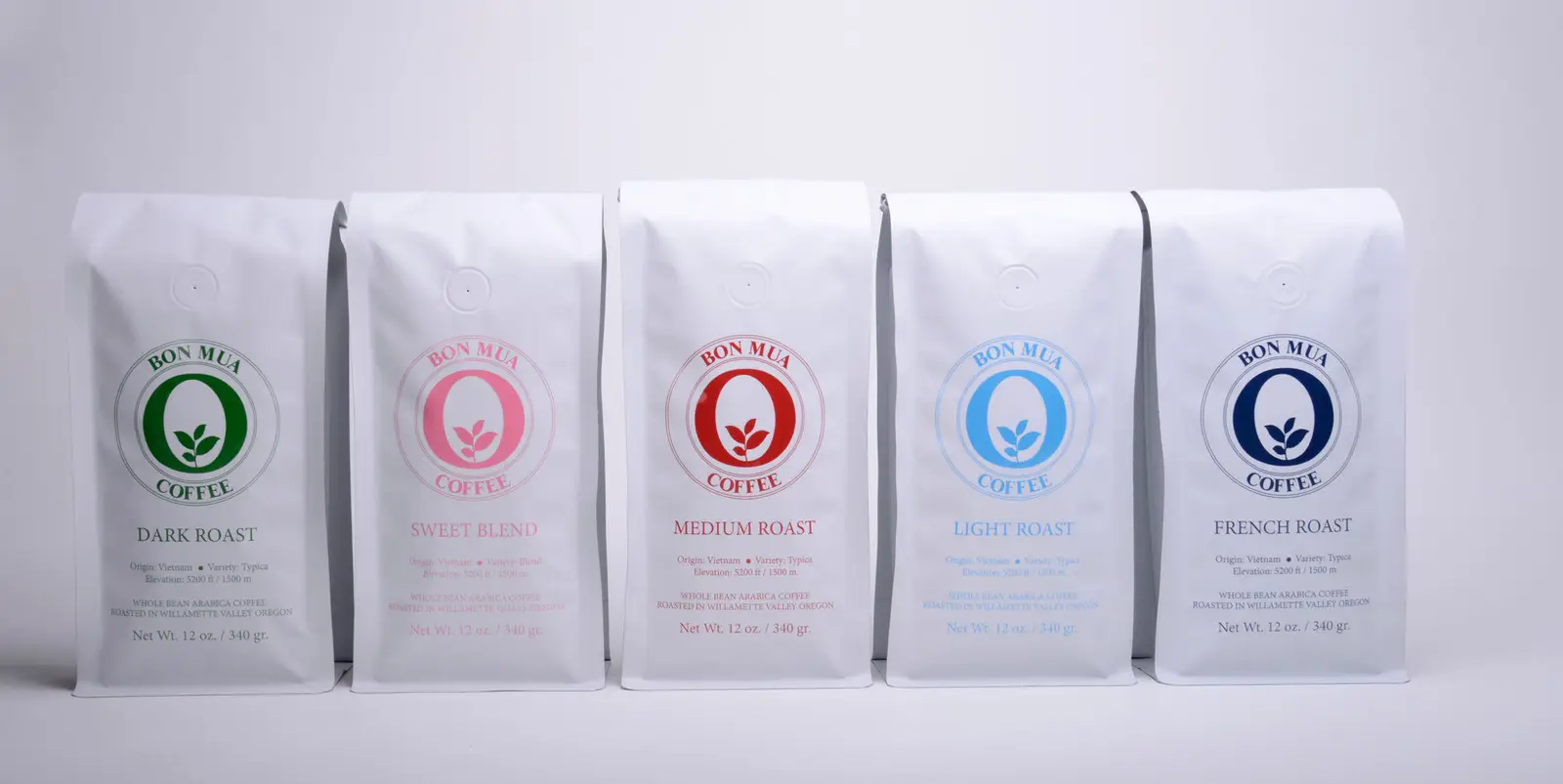 Five white Bon Mua coffee bags standing in a row — Dark, Sweet Blend, Medium, Light and French roasts — each with its colored O-shaped logo, on a pale grey background.