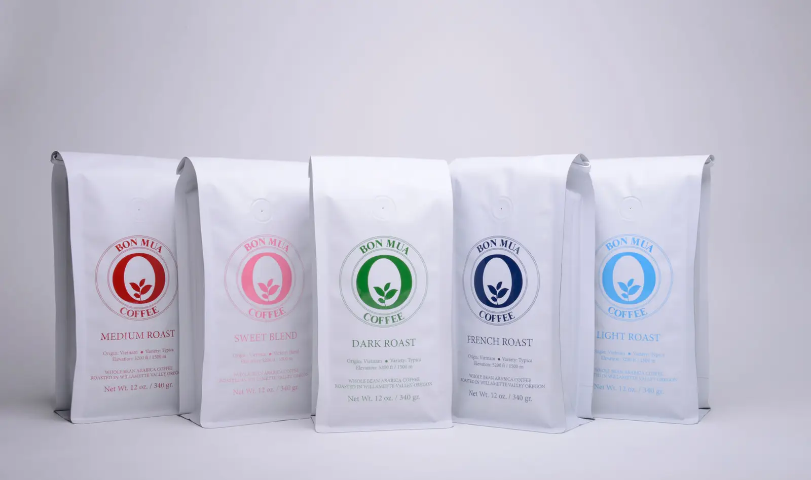 Five white Bon Mua coffee bags lined up in a row on a pale grey background, each printed with a differently colored O-shaped logo — red, pink, green, navy and blue — for the medium, sweet blend, dark, French and light roasts.