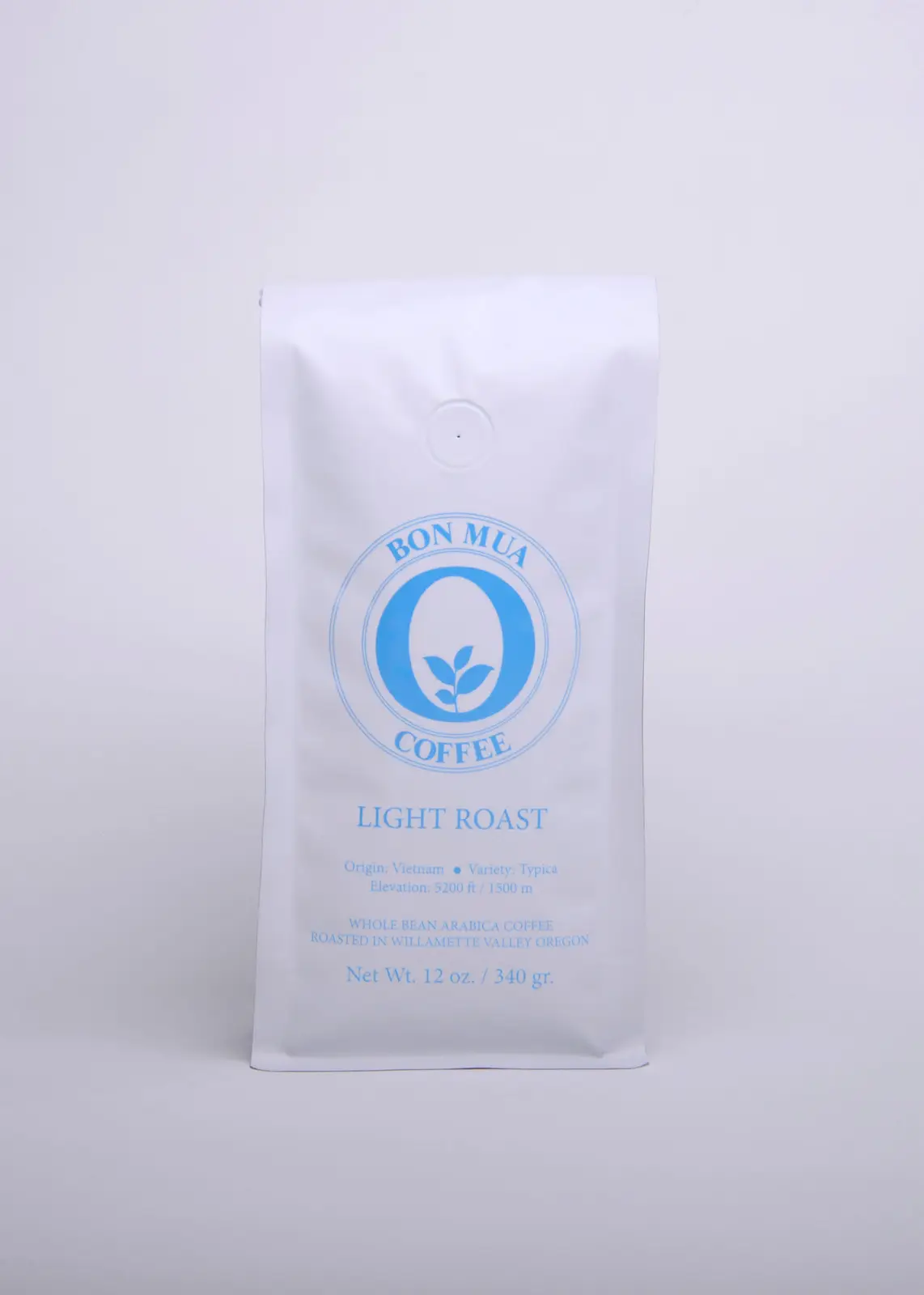 A single white Bon Mua Light Roast coffee bag with a sky-blue O-shaped logo, photographed straight on against a pale grey background.