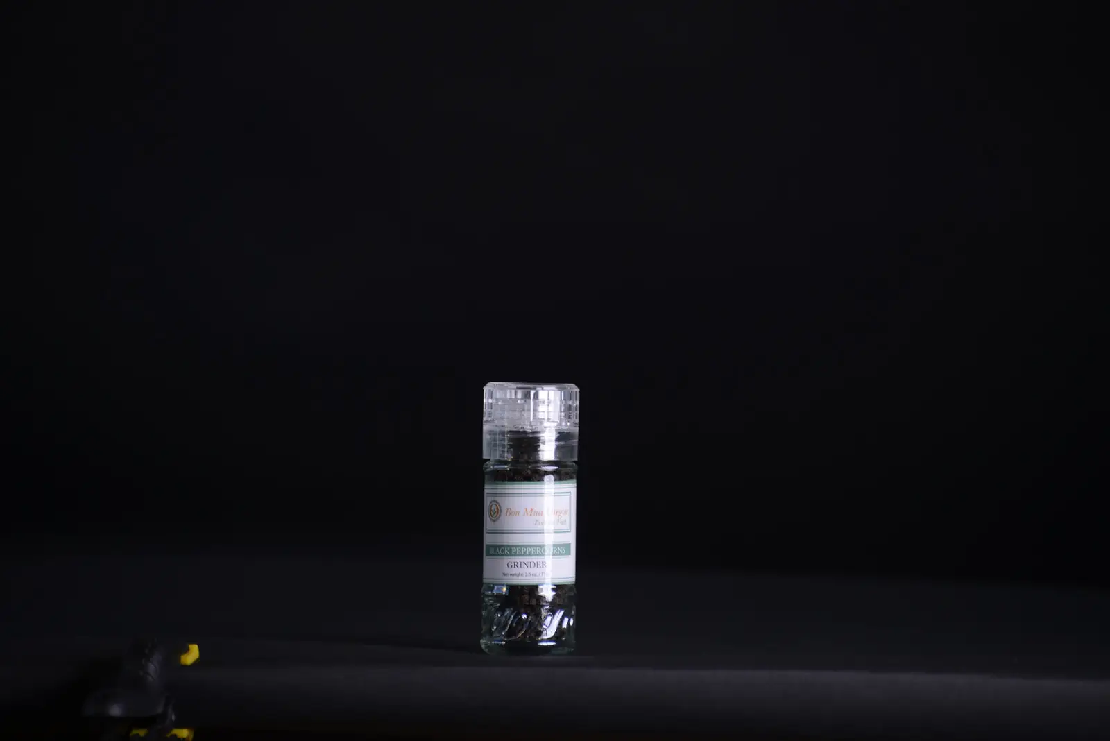 A clear refillable grinder jar of Bon Mua whole black peppercorns with a clear grinder cap, lit against a dark background.