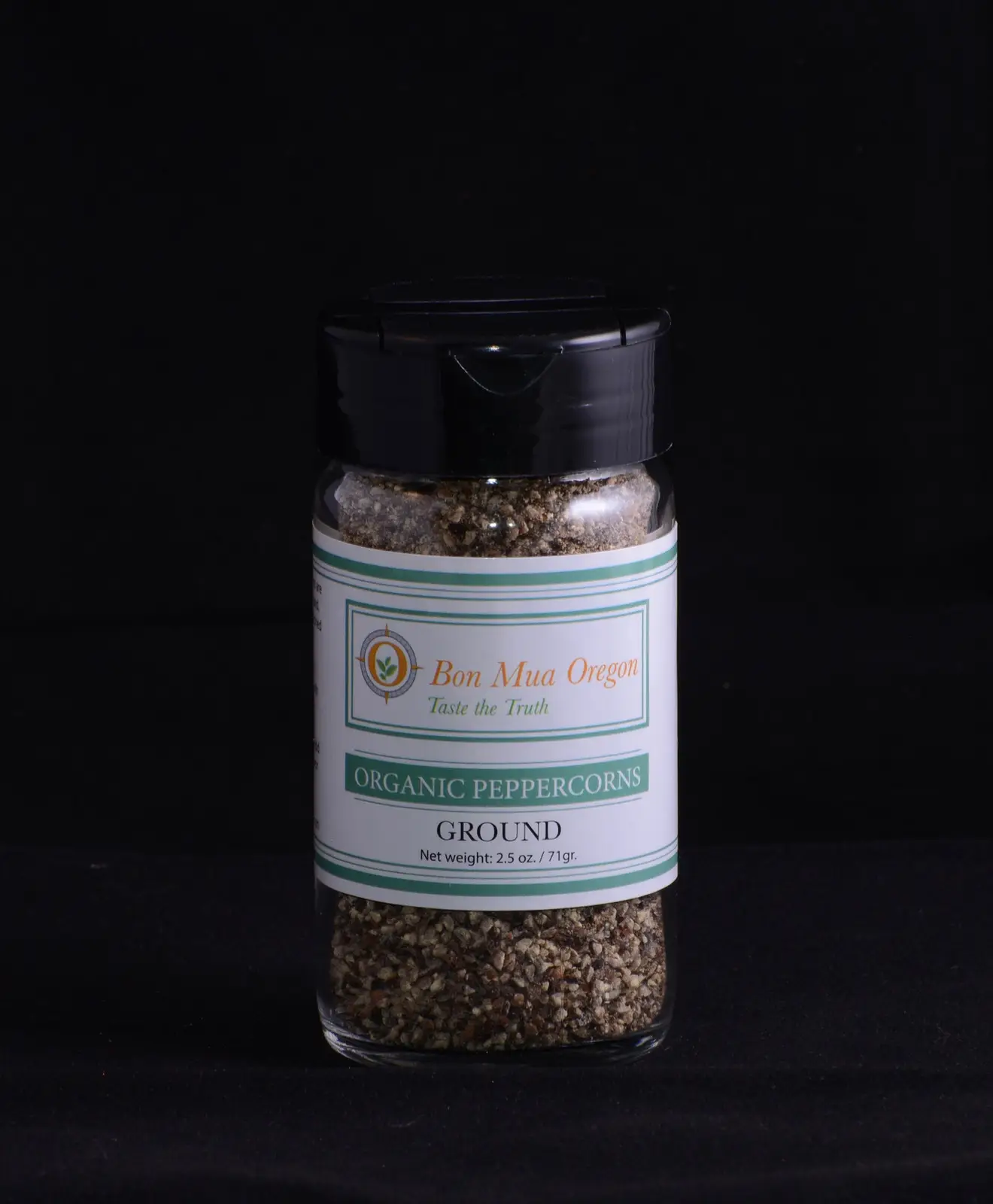 A glass shaker jar of Bon Mua ground black pepper with a black flip-top lid and a green-and-white label, lit against a black background.