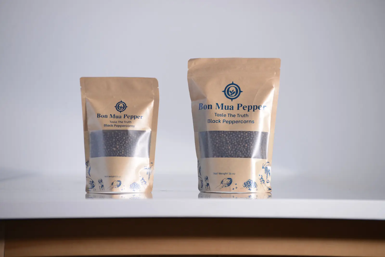 Two kraft-paper stand-up pouches of Bon Mua black peppercorns with clear windows showing the dark whole berries, labeled with a navy compass logo, on a white table.