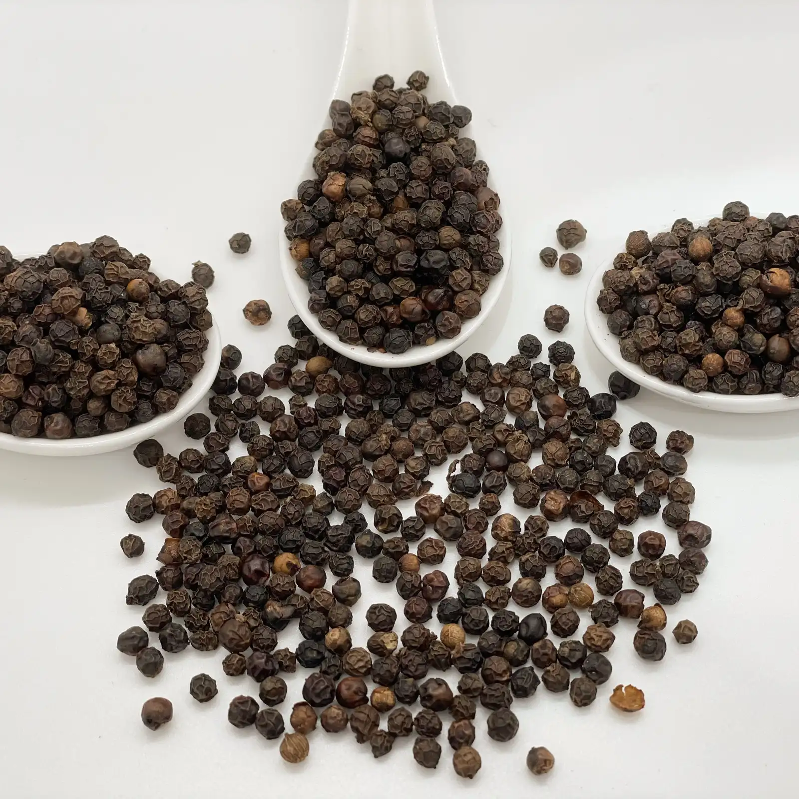 Whole black peppercorns spilling from white ceramic spoons on a pale surface.