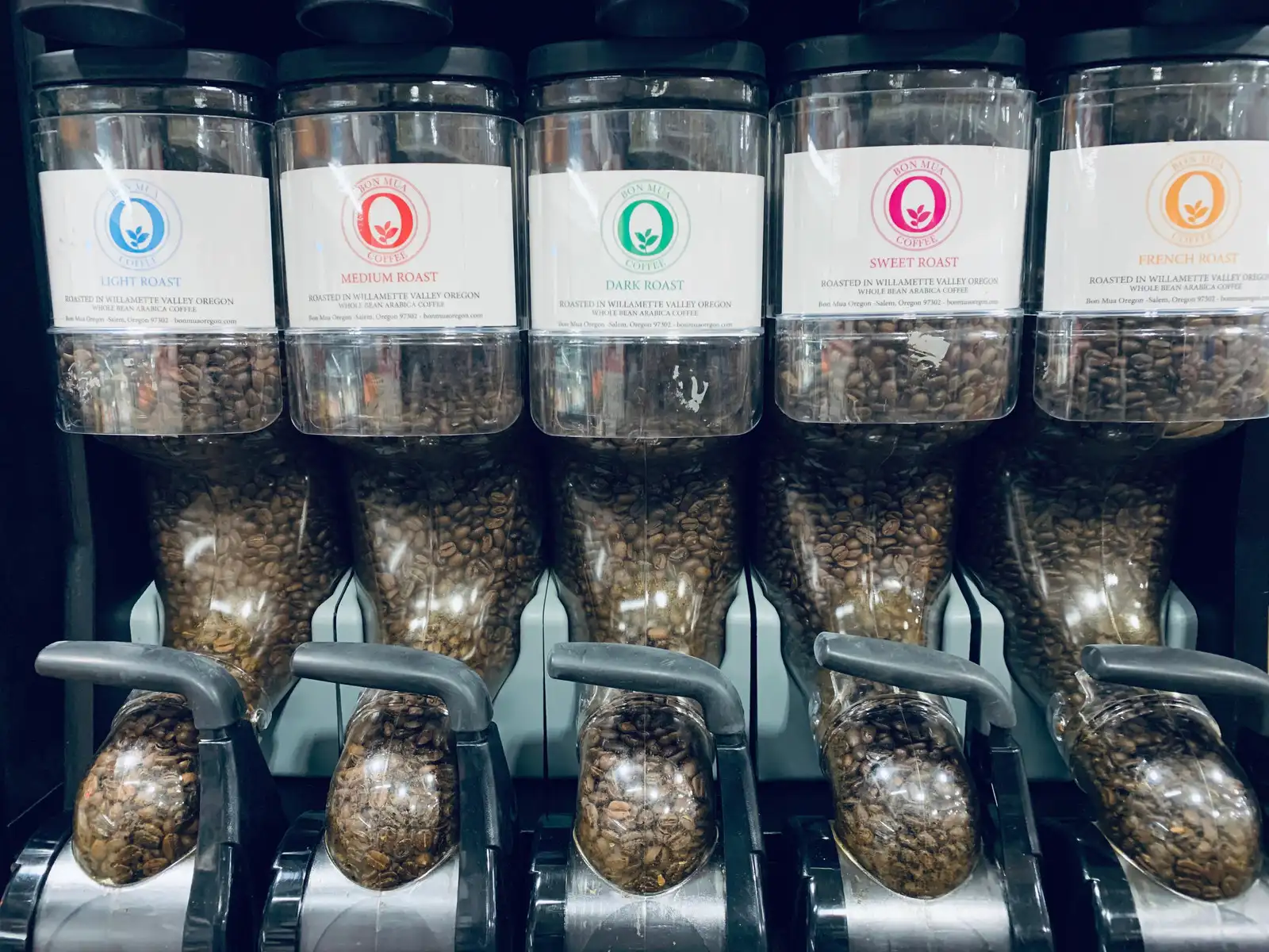 Bulk gravity dispensers filled with Bon Mua whole coffee beans, labeled by roast.