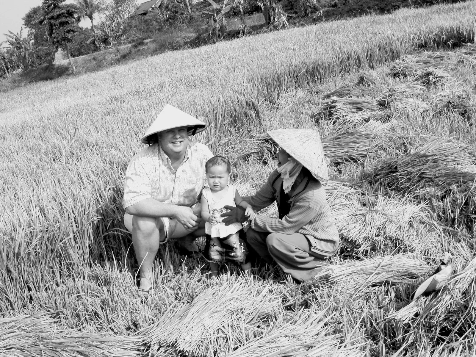 Black-and-white photo of John Marberry and a Vietnamese woman in conical hats crouching with a small child in a freshly cut rice field.