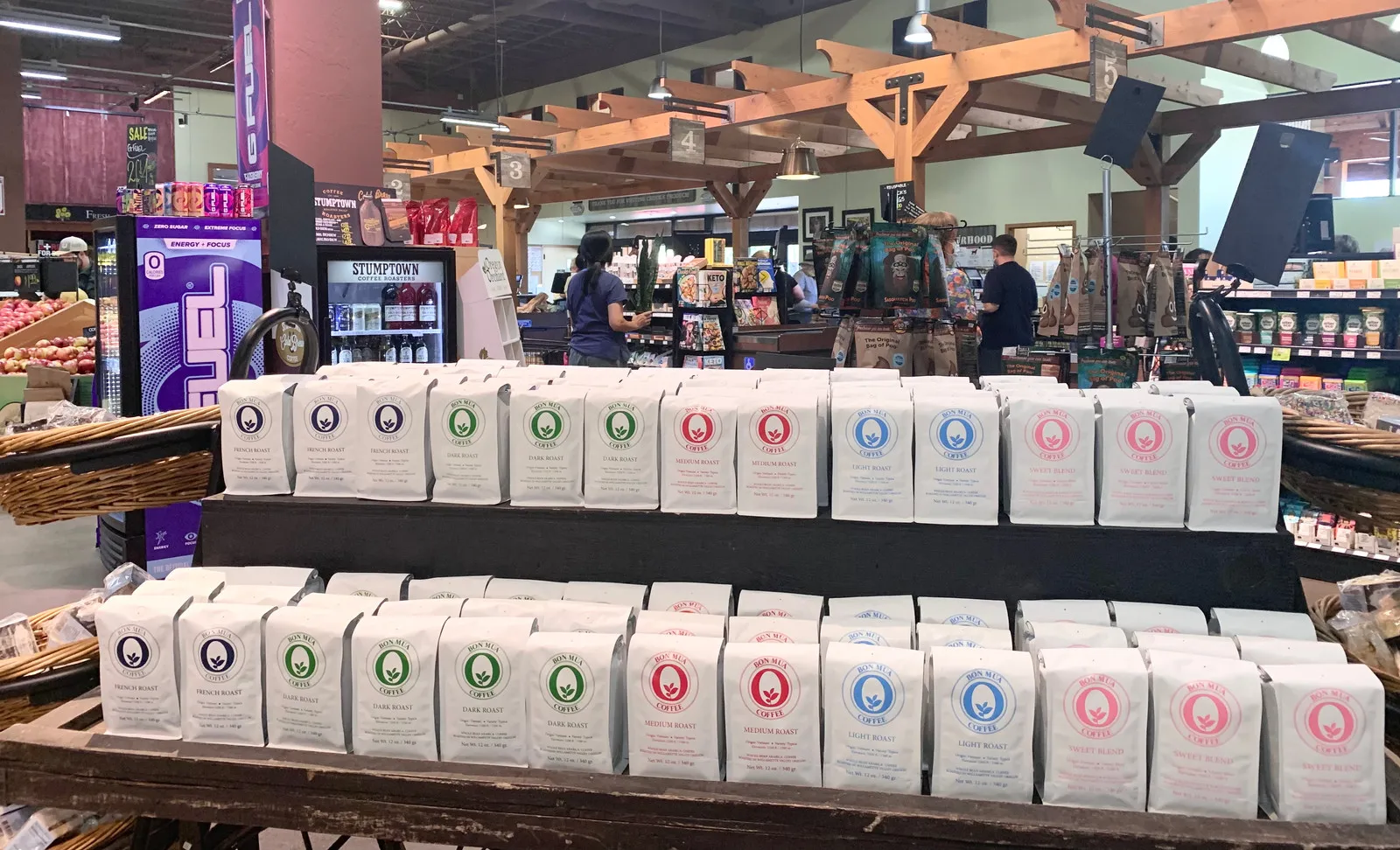 A large table display of dozens of white Bon Mua coffee bags in every roast color inside a wood-beamed grocery store.