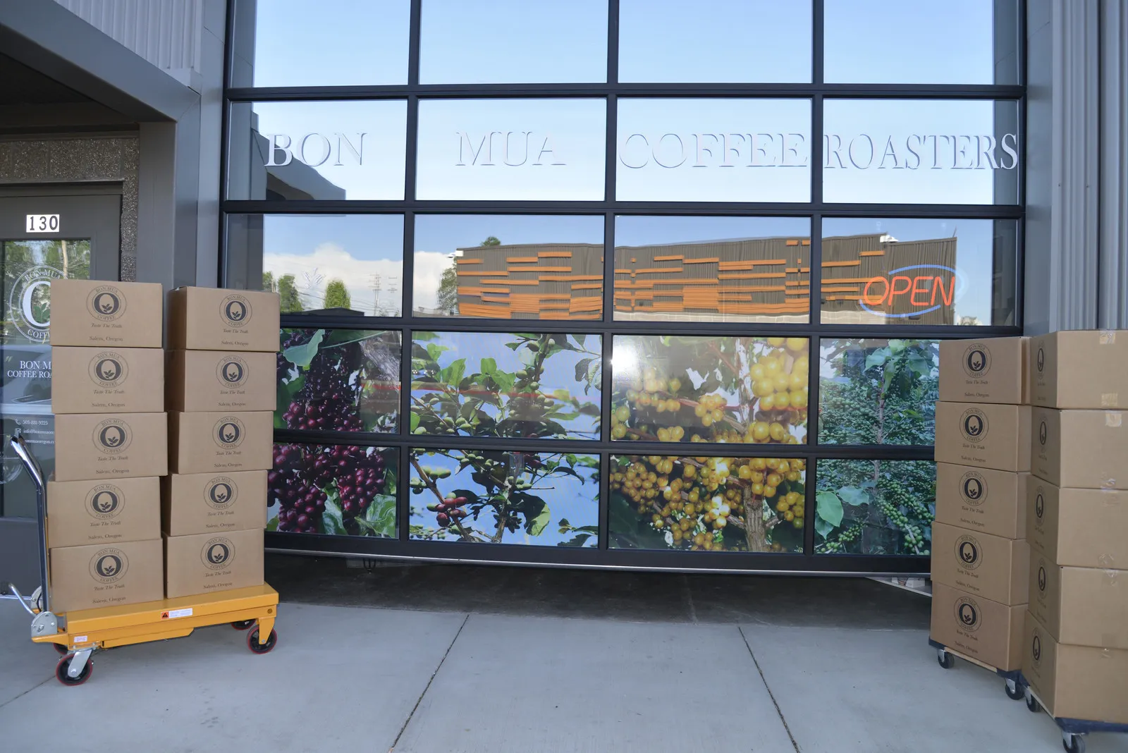 The Bon Mua Coffee Roasters storefront in Salem: a glass garage-door front etched with the company name over a mural of coffee cherries, an OPEN neon sign, and stacked shipping boxes on carts by the door.