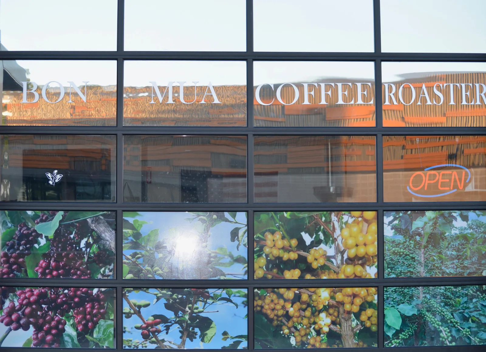 Close view of the Bon Mua Coffee Roasters window with the etched name, a coffee-cherry mural, and an orange OPEN neon sign.