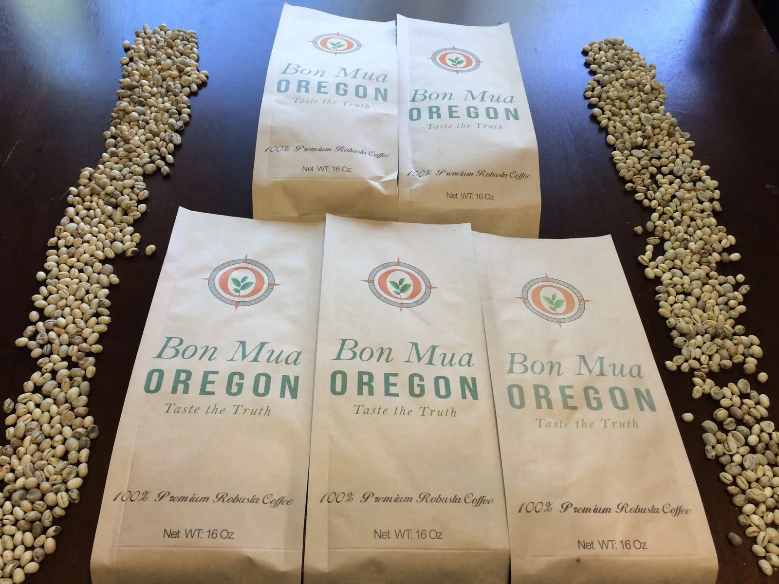 Five kraft-paper bags reading Bon Mua Oregon — Taste the Truth, 100% Premium Robusta Coffee, arranged with lines of green coffee beans on a dark wood table.