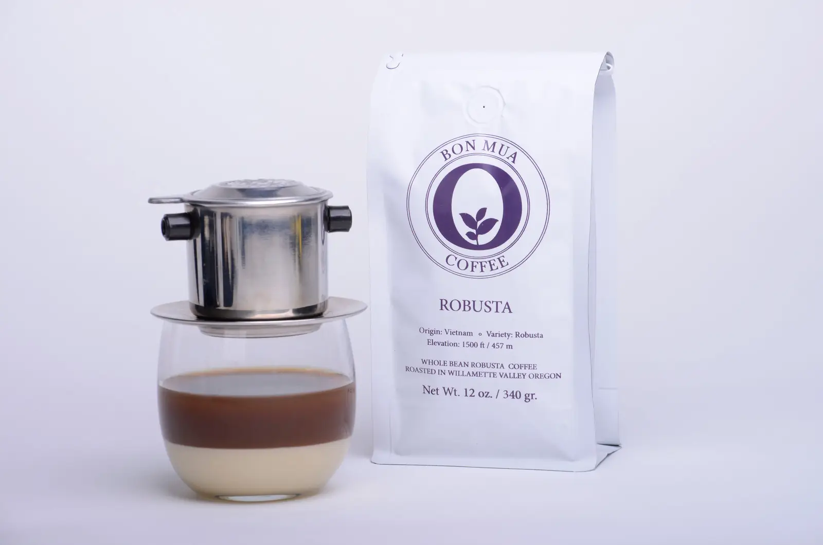 A white Bon Mua Robusta coffee bag with a purple O-shaped logo standing beside a Vietnamese phin filter dripping fresh coffee into a glass over a layer of condensed milk.