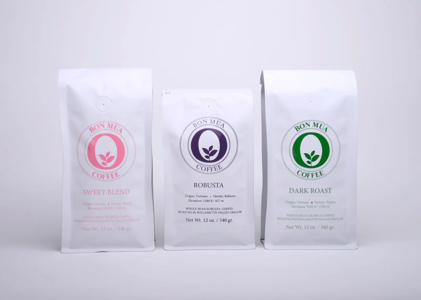 Three white Bon Mua coffee bags standing together — a pink-logo Sweet Blend, a purple-logo Robusta, and a green-logo Dark Roast — on a pale grey background.
